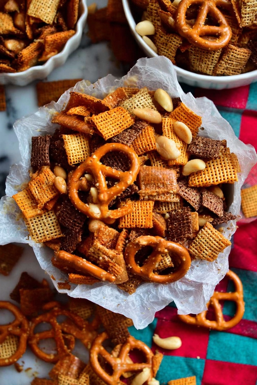 Christmas Eve Savory Snack Mix, savory holiday snack mix, holiday party snack recipes, crowd-pleasing snack mix, festive savory snacks - A close-up of a mix of snacks piled in a bowl lined with white parchment paper, sitting on a white marbled surface; the mix shows layers of light orange square crackers with a smooth texture, dark brown woven cereal pieces with a rough texture, golden pretzels with a shiny surface and salt specks, and small pale yellow peanuts scattered throughout. The background has a colorful checkered cloth with red, turquoise, and beige squares, adding contrast to the snack pile. Some of the snack mix is spilled outside the bowl onto the marbled surface, with two white bowls partially visible containing more of the same snack mix. photo taken with an iphone --ar 2:3 --v 7