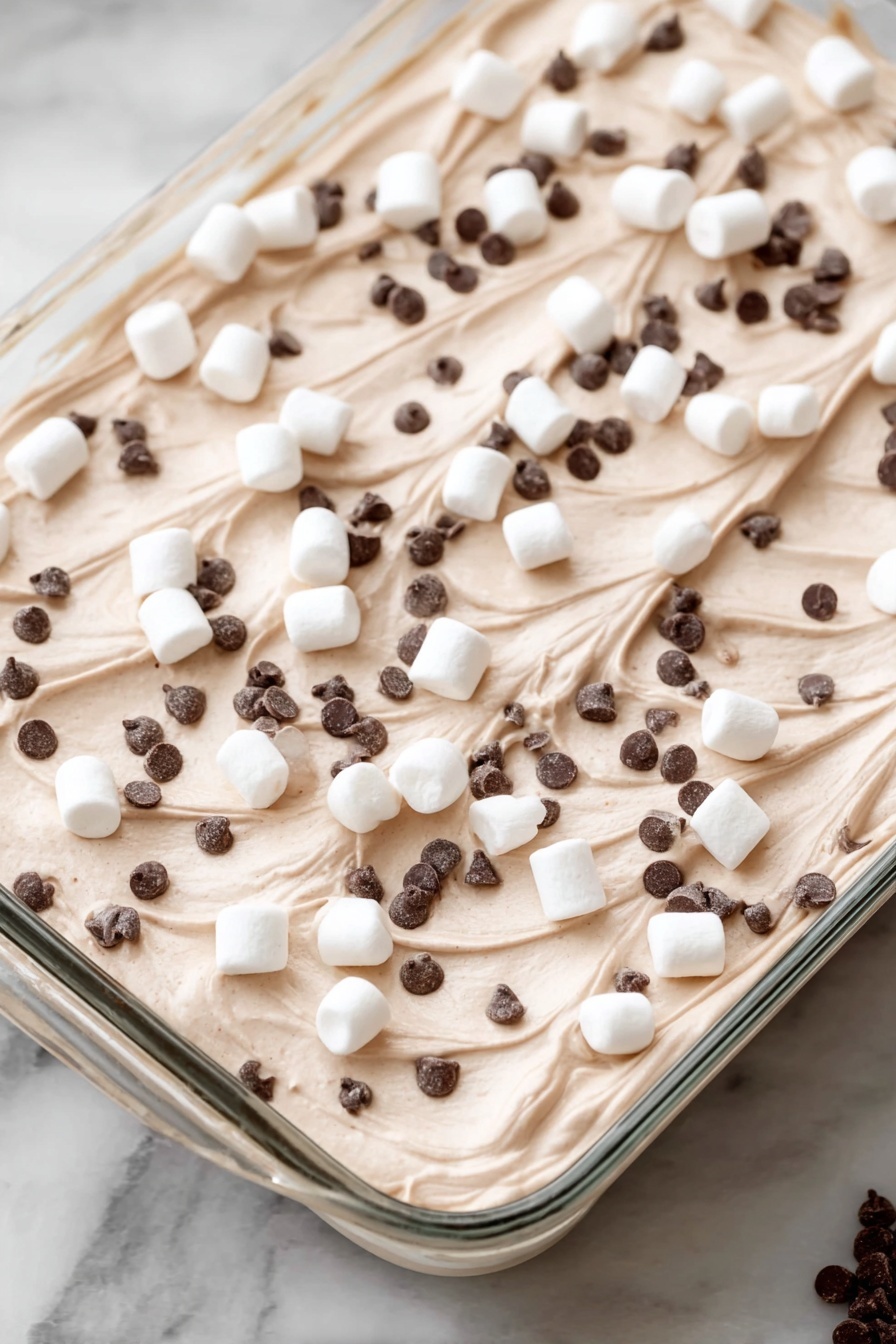 Hot Chocolate Poke Cake, chocolate poke cake, hot cocoa dessert, marshmallow fudge cake, easy chocolate cake recipe - A dessert in a clear rectangular glass dish with one thick layer of light brown creamy mousse or whipped topping, spread evenly with soft swirls on the surface. Scattered on top are many small white marshmallows along with a good amount of very tiny dark brown chocolate chips spread all over. The dish is placed on a white marbled surface. Photo taken with an iphone --ar 2:3 --v 7