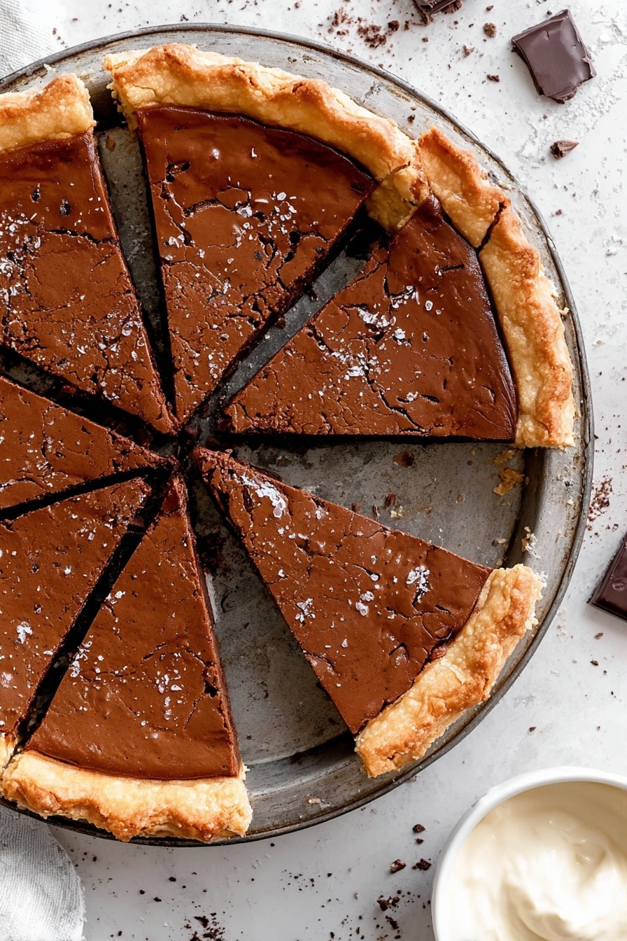 Fudgy Chocolate Brownie Pie, Chocolate Brownie Pie, Best Brownie Pie, Easy Brownie Pie, Chocolate Dessert - The image shows a round pie cut into six slices. The pie has a thick, shiny, dark brown top layer with a cracked texture and a golden, flaky crust around the edges. The pie rests on a metal tray with a dark, worn finish. The background is a white marbled surface with scattered chocolate bits. At the bottom right corner, there's a small white bowl filled with smooth, light cream. photo taken with an iphone --ar 2:3 --v 7