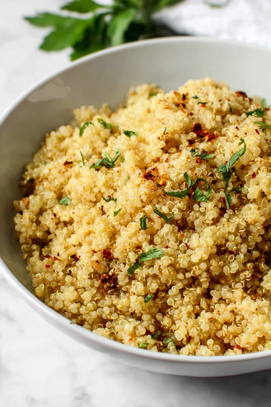 Garlic Butter Quinoa, flavorful quinoa side dish, easy garlic butter quinoa, quick quinoa recipes, best side dishes with quinoa - A close-up view of a white bowl filled with cooked quinoa, showing light yellow grains mixed with small green herb pieces sprinkled on top, along with a few red spice flakes and some black pepper. A golden spoon is placed inside the bowl on the right side. The bowl sits on a white marbled surface with some green herb leaves blurred in the background. photo taken with an iphone --ar 2:3 --v 7