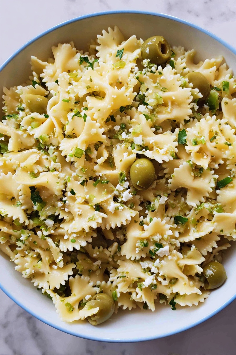 Dirty Martini Pasta Salad, easy pasta salad recipes, flavorful pasta salad ideas, briny pasta salad, party pasta salads - A close-up view of a large white bowl filled with curly-edged pasta that is light yellow in color, mixed evenly with bright green sliced olives and sprinkled with small bits of light green herbs and crumbled white cheese. The pasta is separated nicely, showing the wavy edges and soft texture, while the green olives add contrast throughout. The bowl sits on a white marbled surface, creating a clean and fresh look. photo taken with an iphone --ar 2:3 --v 7