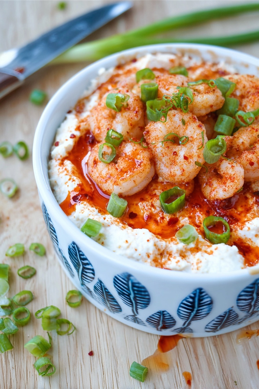 Shrimp Cocktail Dip, seafood appetizer recipes, easy party dips, creamy shrimp dip, quick appetizer ideas - The dish shows three layers in a white bowl with blue leaf patterns. The bottom layer is a creamy white spread with a smooth texture. The middle layer is a bright red sauce that looks thick and slightly shiny. The top layer consists of small cooked shrimp, seasoned with red spices giving them a light orange color, and sprinkled with chopped green onions for a fresh green contrast. The bowl is placed on a light wooden surface with a few green onion pieces scattered around and a knife with some sauce on the blade nearby. Photo taken with an iphone --ar 2:3 --v 7