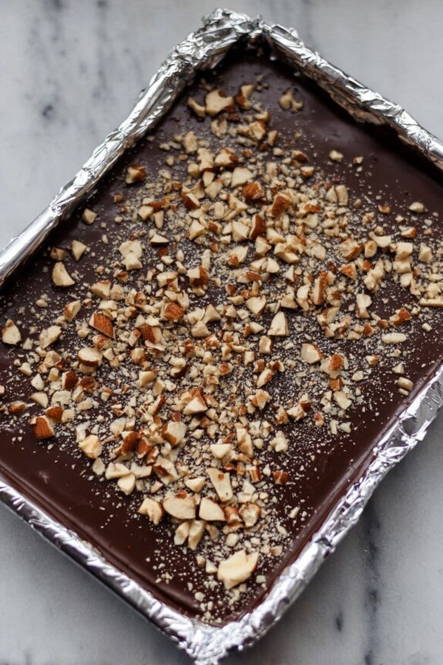 English Toffee with Chocolate and Nuts, best English Toffee, homemade toffee recipe, easy toffee with nuts and chocolate, crunchy buttery toffee - This image shows a foil-lined rectangular tray filled with a smooth, dark chocolate layer. On top of the chocolate is an even spread of chopped nuts, both larger and smaller pieces, creating a textured surface. The foil edges around the tray are crinkled and shiny, reflecting light softly. The background is a white marbled texture. photo taken with an iphone --ar 2:3 --v 7