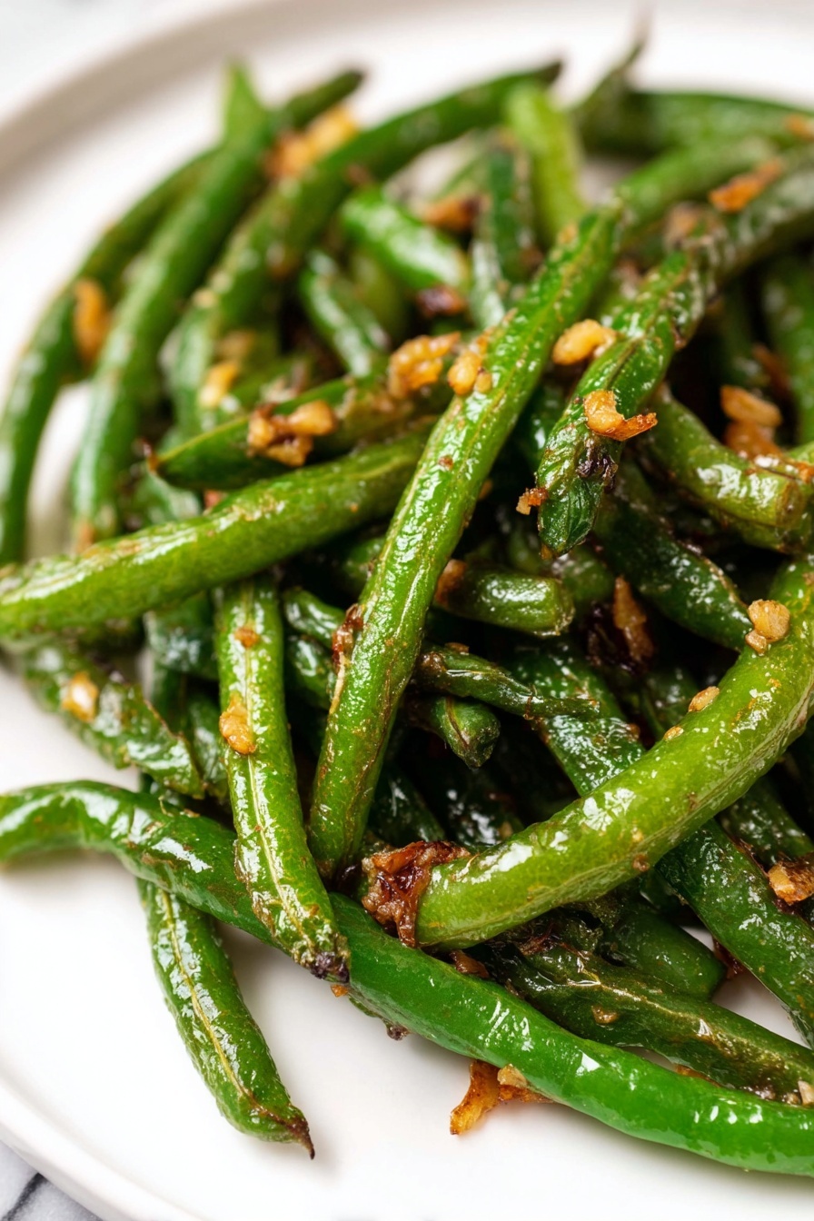 Garlic Green Beans Stir Fry, healthy green bean side dish, quick vegetable stir fry, garlic green bean recipe, easy green beans recipe - A pile of cooked green beans with browned, slightly wrinkled skin is placed on a white paper towel over a white marbled surface. To the upper left of the green beans, there is a clear round glass bowl filled with a light brown sauce mixed with small pieces of chopped garlic or onion. The sauce has a glossy texture. The background is a white cloth that partially covers the surface. photo taken with an iphone --ar 2:3 --v 7