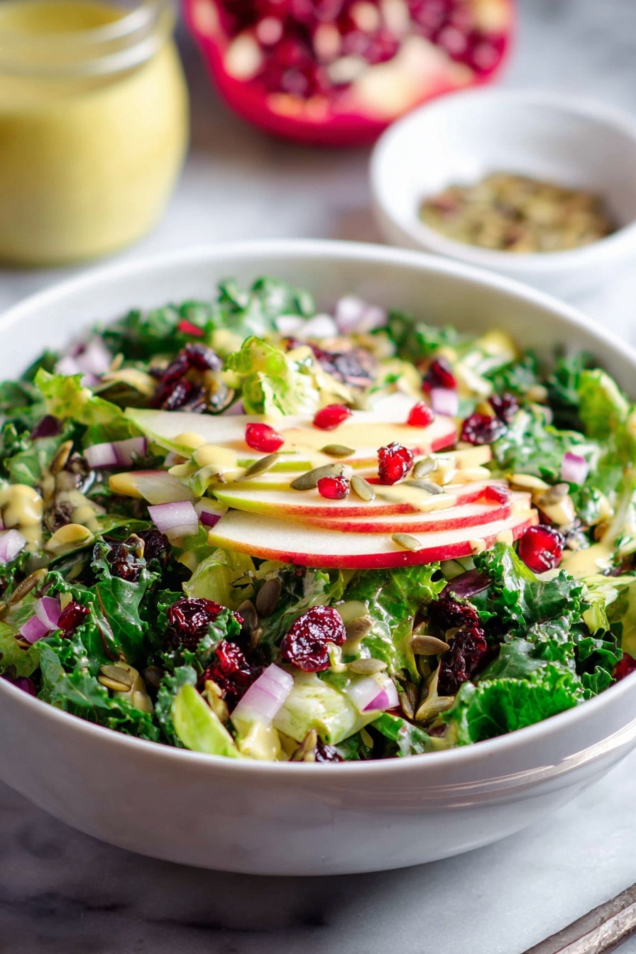 Shaved Brussels Sprouts with Pomegranate, Brussels Sprouts Salad with Pomegranate, Healthy Brussels Sprouts Salad, Christmas Brussels Sprouts Salad, Easy Brussels Sprouts Salad - A white bowl is filled with a fresh salad showing multiple layers: at the bottom are bright green leafy vegetables, followed by thin slices of red and green apple. On top, there are pieces of chopped red onion, dark green kale leaves, bright red pomegranate seeds, dried red cranberries, and scattered sunflower seeds, all mixed together. The salad is lightly dressed with a pale yellow dressing that adds a slight shine to the ingredients. The bowl is placed on a white marbled surface, with a blurred jar of light yellow dressing and a small white bowl of seeds in the background. photo taken with an iphone --ar 2:3 --v 7