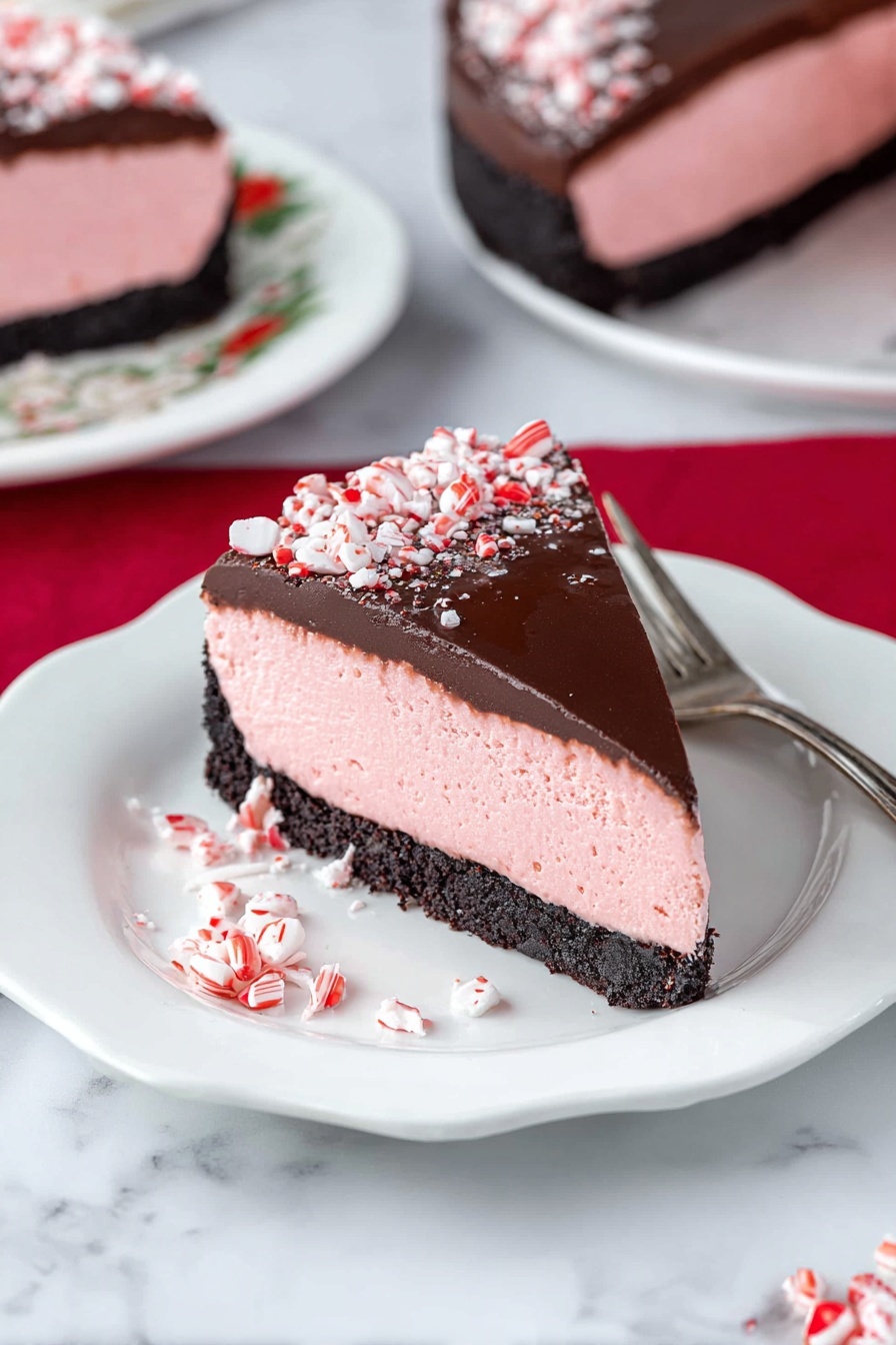 Peppermint Cheesecake with Chocolate Ganache, festive peppermint cheesecake, holiday dessert recipes, mint chocolate cheesecake, easy peppermint dessert - A slice of dessert with three distinct layers sits on a white plate with scalloped edges; the bottom layer is a dark, crumbly crust, the middle layer is a thick, smooth pink mousse, and the top layer is a glossy dark chocolate coating sprinkled with crushed red-and-white candy pieces. The slice is garnished with more crushed candies at the base and is accompanied by a silver fork placed nearby. In the background, parts of the full dessert and another slice on a white plate with floral patterns rest on a white marbled surface, accented by a red cloth. Photo taken with an iphone --ar 2:3 --v 7