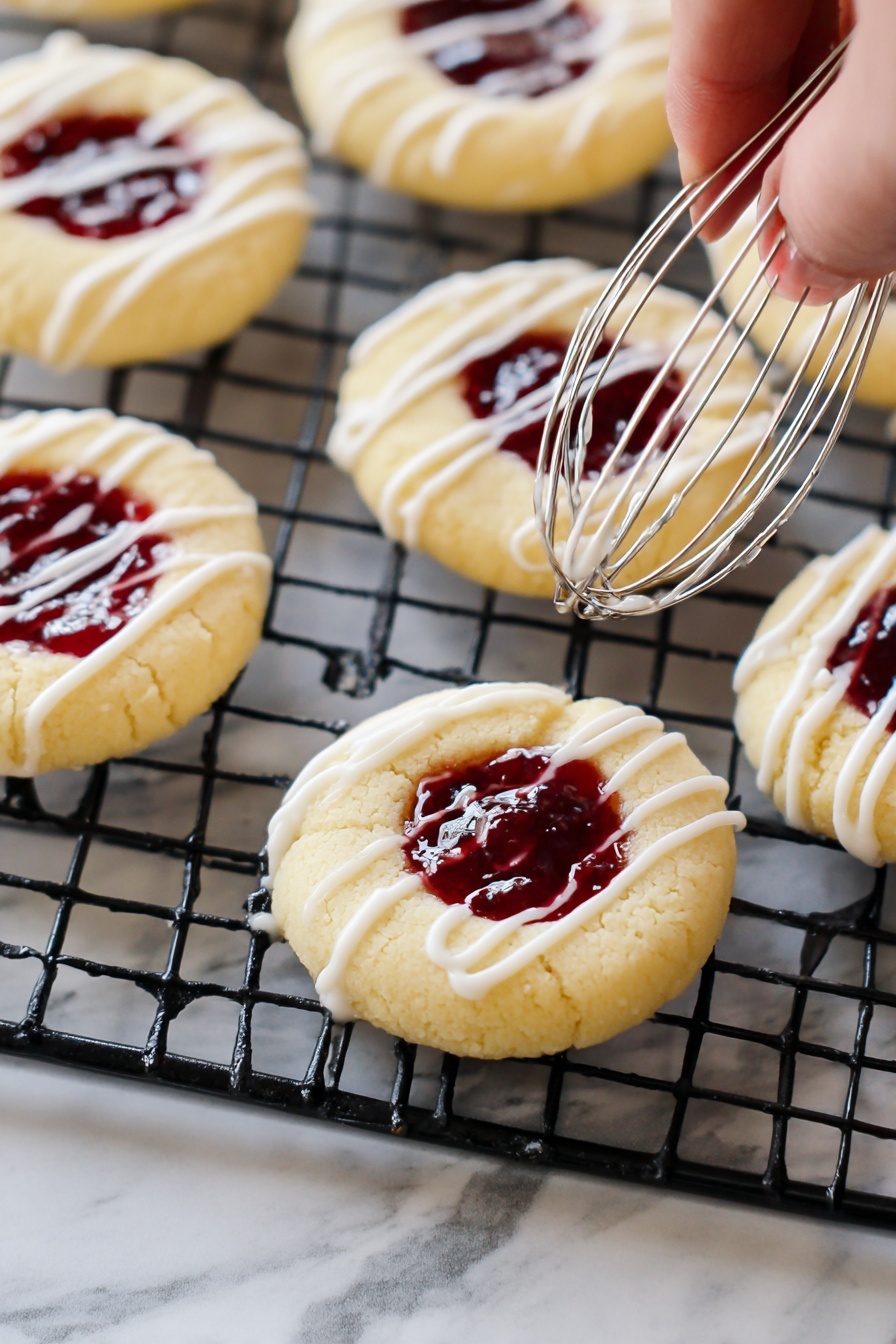 Raspberry Thumbprint Cookies, Raspberry jam cookies, buttery thumbprint cookies, fruit-filled cookie recipes, easy holiday cookies - The image shows round cookies with a thumbprint center filled with dark red jam, placed on a black cooling rack over a white marbled surface. Each cookie has a pale yellow dough base with a smooth texture. White icing is being drizzled in thin stripes over the cookies by a metal whisk held by a woman's hand from the right side of the image. The jam in the cookie centers has a glossy surface, creating a shiny contrast with the soft, matte cookie dough. Photo taken with an iphone --ar 2:3 --v 7