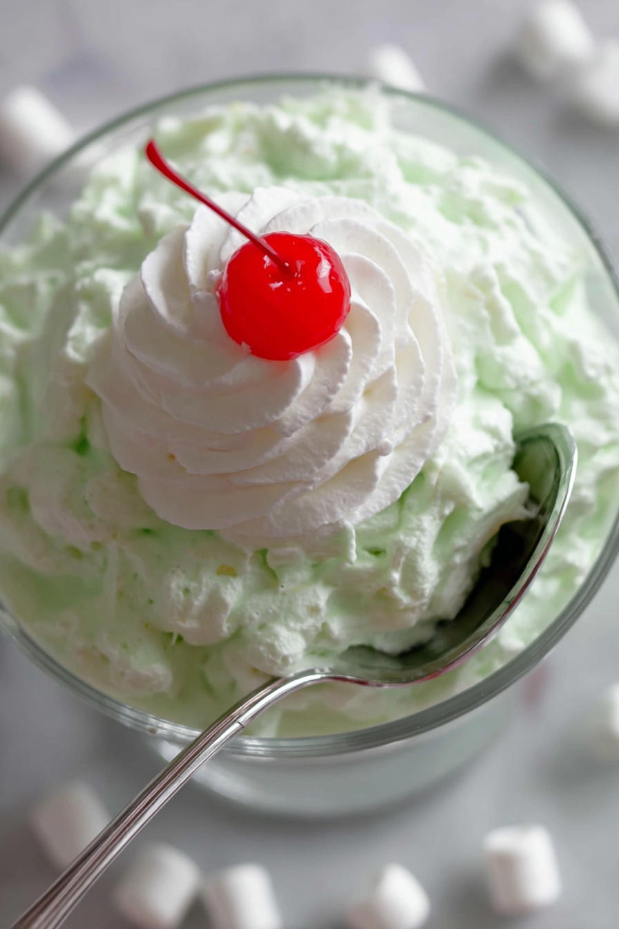 Easy Watergate Salad with Pineapple and Pecans, Watergate Salad recipe, quick fruity dessert, no-cook pineapple salad, nostalgic party side dish - A close-up image of a layered dessert in a clear glass bowl placed on a white marbled surface with mini white marshmallows scattered around. The bottom layer is a creamy, light green textured mixture that looks fluffy and thick. On top of this green layer is a smooth, white dollop of whipped cream shaped in a spiral pattern. Sitting right in the center of the whipped cream is a bright red, shiny cherry. A silver spoon is partly inserted into the green layer, curving around the side of the bowl. photo taken with an iphone --ar 2:3 --v 7