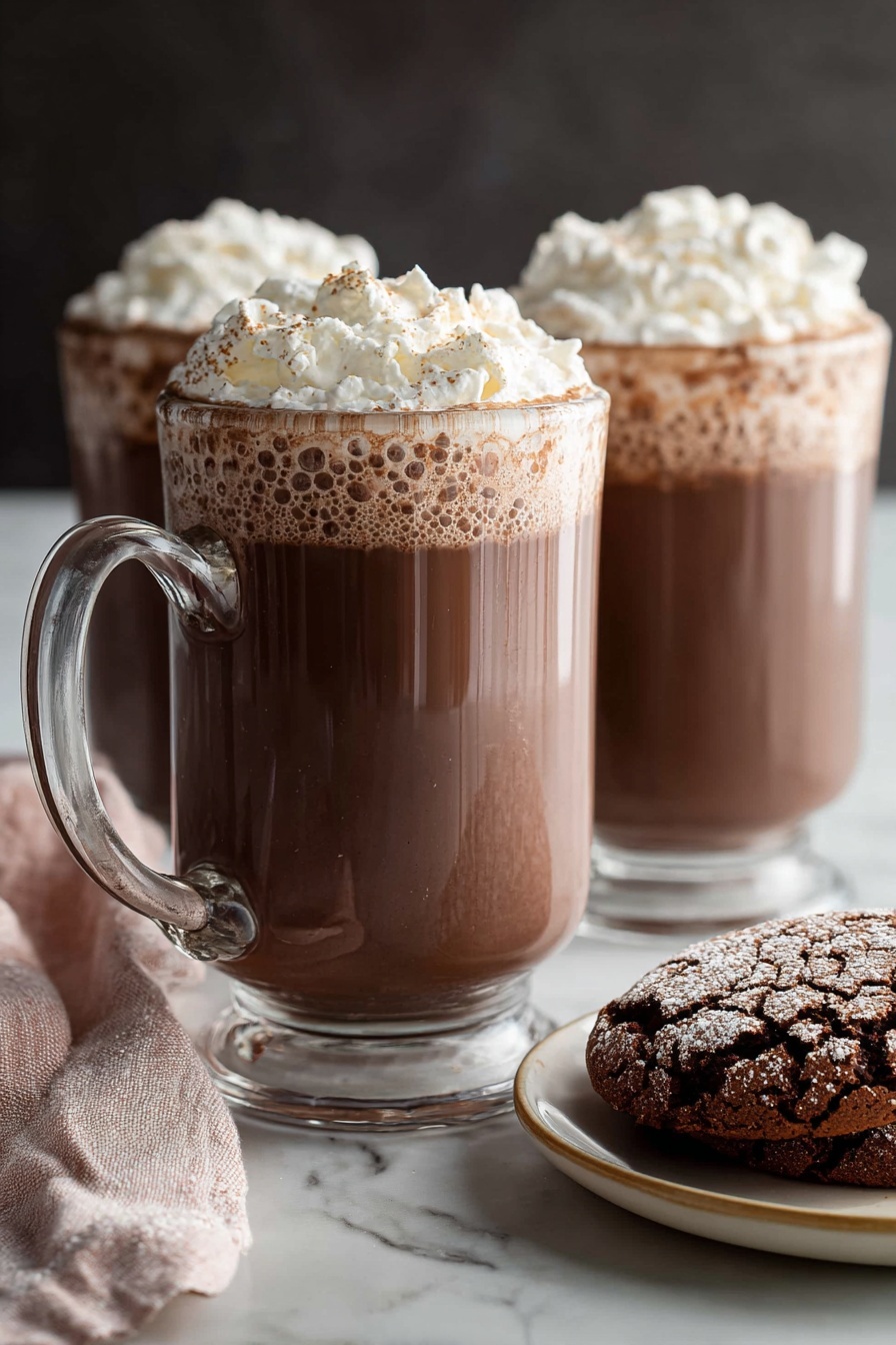 Creamy Homemade Hot Chocolate, homemade hot chocolate, easy hot chocolate recipe, rich hot chocolate, cozy winter drinks - Two clear glass mugs filled with rich brown hot chocolate, topped with a thick layer of light white whipped cream that has a fluffy texture and soft peaks. The mugs have sturdy handles positioned on the left, and the hot chocolate shows slight bubbles near the edges under the whipped cream. In the foreground, on the right side, there is a white plate with a beige rim holding two cracked chocolate cookies with a powdery surface. A soft pink and white cloth napkin is casually placed on the left side near the front mug. All items sit on a smooth white marbled surface with a dark, blurred background behind them. Photo taken with an iphone --ar 2:3 --v 7