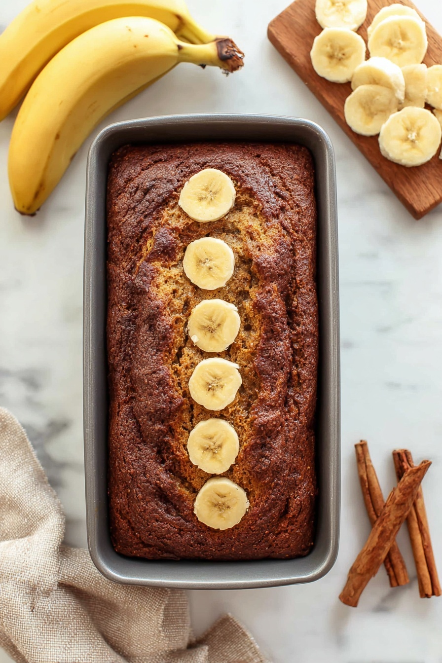 Cinnamon Banana Bread with Swirl, banana bread with cinnamon swirl, cinnamon banana loaf, moist banana bread recipe, easy cinnamon swirl bread - A thick rectangular loaf of dark brown banana bread rests in a grey baking pan, with five round banana slices lined up in the center top of the bread. To the left, two whole yellow bananas lay curved on a white marbled surface. To the right, a wooden board holds several light yellow banana slices arranged in overlapping circles. At the bottom right corner, three brown cinnamon sticks rest on the white marbled surface, and a beige cloth is partially visible at the bottom left corner. photo taken with an iphone --ar 2:3 --v 7