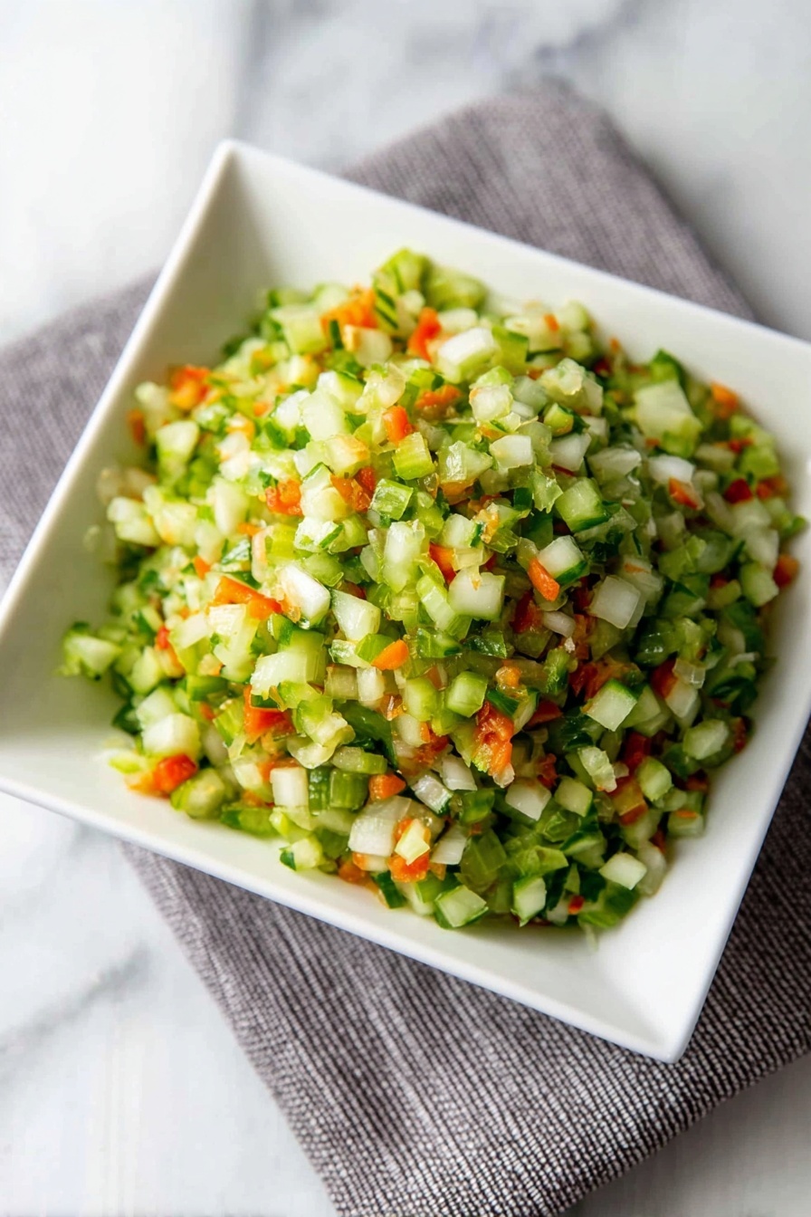 Dill Pickle Salsa, easy pickle salsa, fresh salsa recipes, tangy pickle dip, crunchy cucumber salsa - A close-up view of finely chopped vegetables in a white square bowl placed on a grey cloth on a white marbled surface. The mix has small pieces with mostly green colors from celery or cucumber, bright orange bits from carrots, and some white pieces from onions, creating a fresh and colorful look. The texture appears crisp and moist, with the small vegetable pieces piled up in layers that give a sense of volume and freshness. photo taken with an iphone --ar 2:3 --v 7