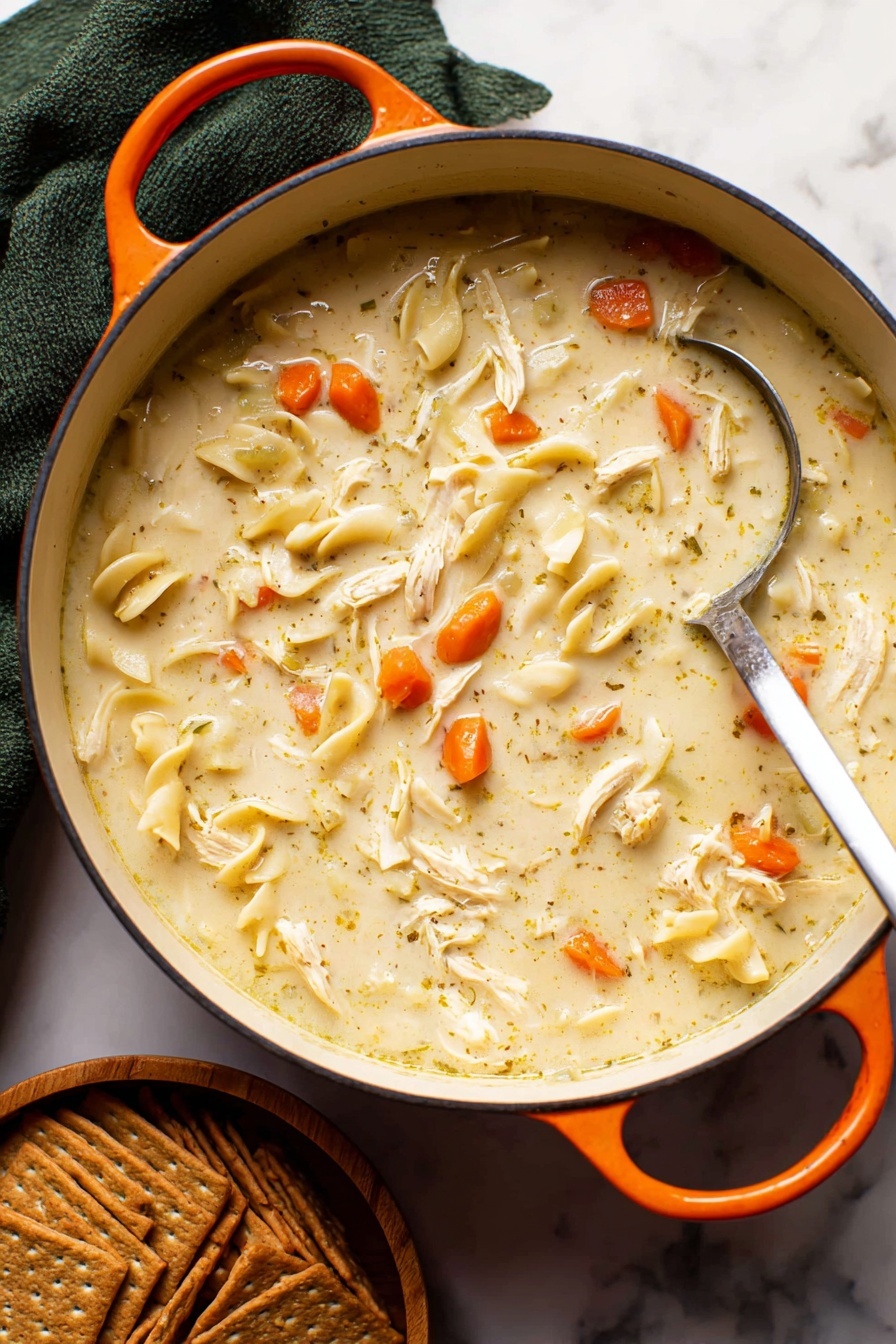 Creamy Chicken Noodle Soup, creamy chicken noodle soup, comforting chicken noodle soup, easy chicken noodle soup, hearty creamy soup - A close-up of a white pot filled with creamy chicken noodle soup, showing a thick light beige broth mixed with small bright orange carrot slices, soft shredded white chicken pieces, and flat light yellowish noodles scattered throughout. A silver ladle is partially dipped in the soup, resting on the right side of the pot. The pot has an orange handle on each side and sits on a white marbled surface. Below the pot, there is a stack of brown crackers arranged neatly in a small wooden bowl. A dark green cloth is folded behind the pot, adding contrast to the scene. Photo taken with an iphone --ar 2:3 --v 7