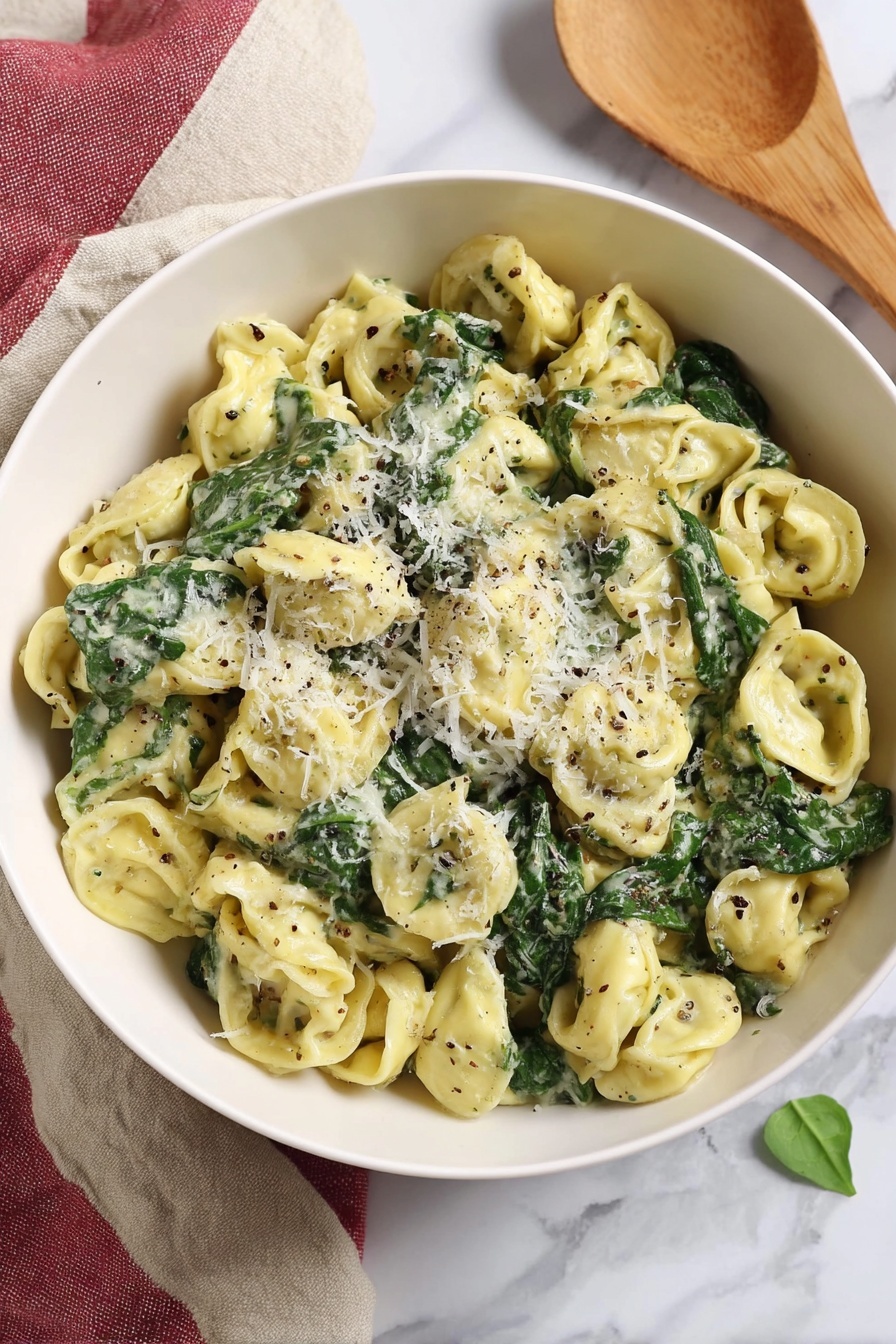 Pesto Tortellini, quick tortellini recipes, easy pasta dinners, cheesy tortellini with pesto, simple weeknight meals - A white bowl filled with three layers of tortellini pasta that is light yellow, mixed with dark green spinach leaves, and coated in a creamy sauce with flecks of black pepper and herbs. On top, there is a light sprinkle of grated white cheese finely covering some pasta and spinach. The bowl sits on a white marbled surface with a beige and red cloth folded beside it, and a wooden spoon rests nearby, all softly lit in natural light. Photo taken with an iphone --ar 2:3 --v 7