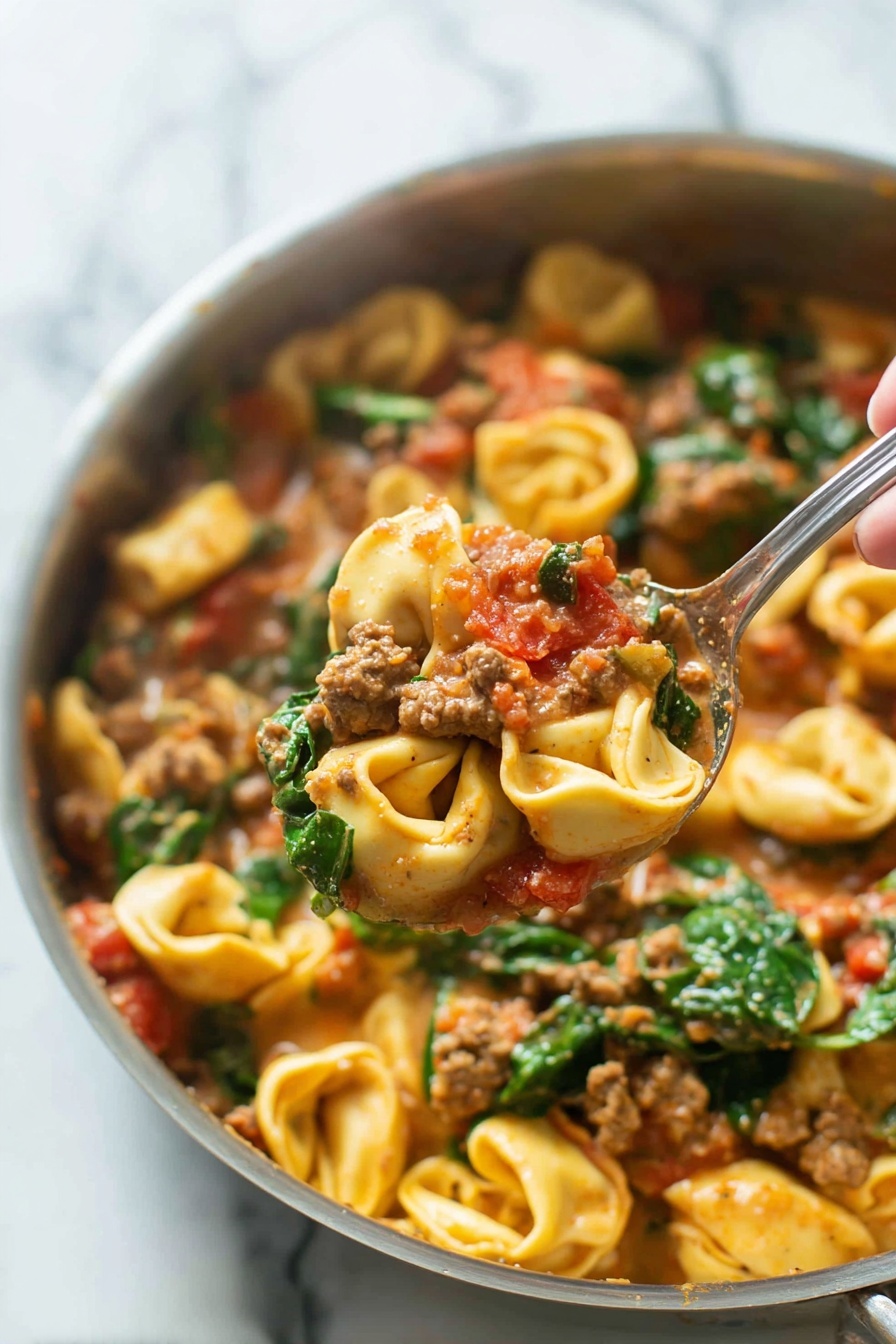 One-Pan Italian Sausage Tortellini, Italian Sausage Tortellini Recipe, Creamy Tomato Sausage Pasta, One-Pan Pasta Dinner, Easy Sausage Tortellini - A close-up image of a cooked pasta dish in a silver pan shows three main layers. The bottom layer is pasta tortellini, small ring-shaped with light yellow color and slightly ruffled edges. The middle layer features ground meat chunks mixed with pieces of red tomato, all coated in a creamy, light brown sauce. The top layer has fresh green spinach leaves scattered throughout, adding vibrant color and texture. A woman's hand holding a silver spoon lifts a portion of the pasta, meat, tomato, and spinach mixture above the pan. The background is a white marbled surface. photo taken with an iphone --ar 2:3 --v 7