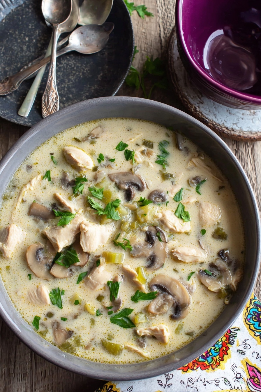 Creamy Chicken Mushroom Soup, comforting chicken and mushroom soup, velvety chicken mushroom soup, easy chicken mushroom soup recipe, hearty mushroom soup - A gray bowl filled with creamy soup that has chunks of light beige cooked chicken, sliced brown and white mushrooms, and small pieces of green celery, all mixed in a pale, creamy broth with green parsley leaves scattered on top, sitting on a wooden table next to a dark gray plate with two silver spoons and a purple bowl, with a white napkin that has colorful patterns to the side, the surface beneath is a white marbled texture photo taken with an iphone --ar 2:3 --v 7