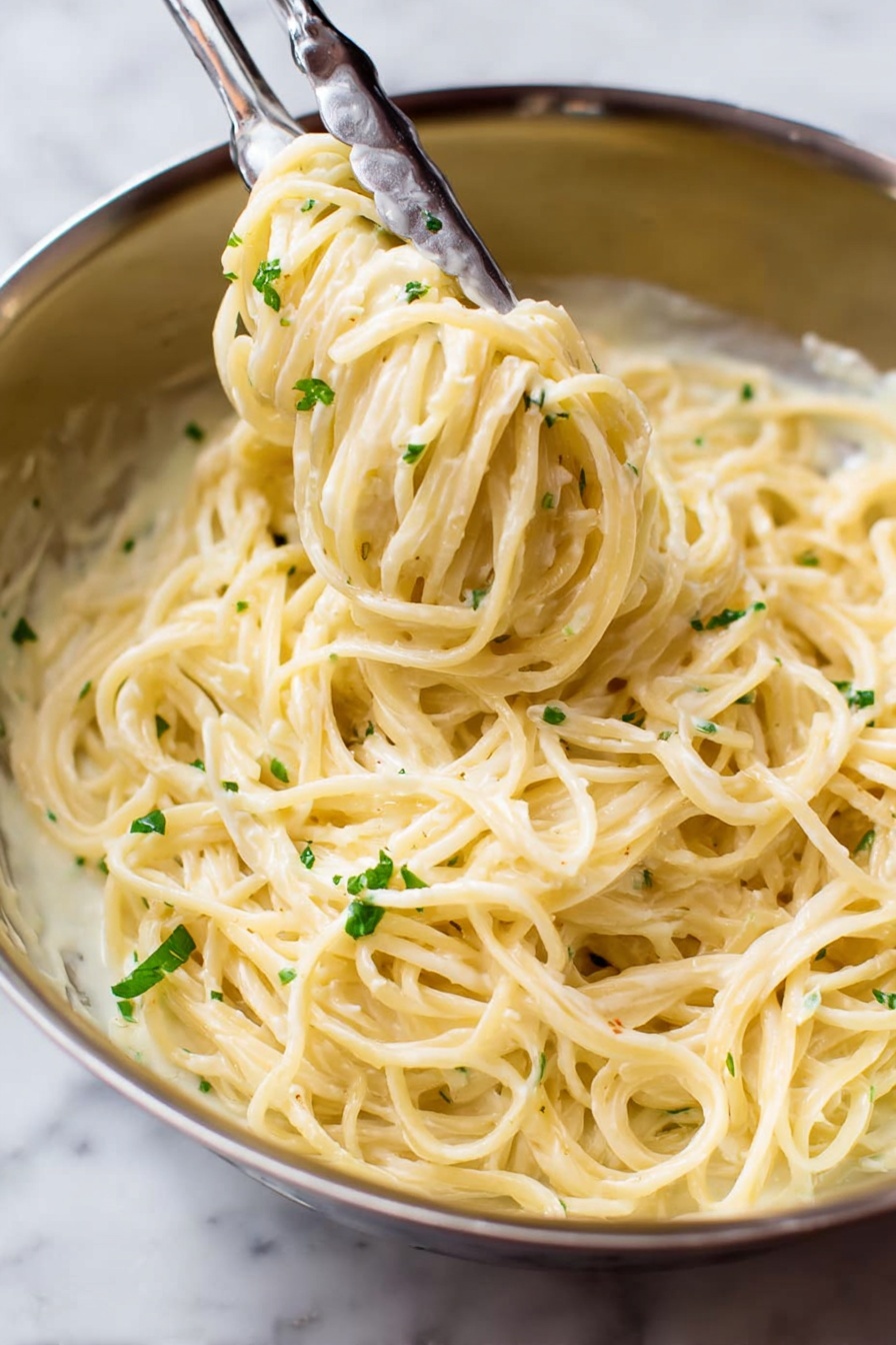 Garlic Parmesan Angel Hair Pasta, quick garlic Parmesan pasta, creamy angel hair pasta, easy one-pot pasta, buttery garlic pasta - A close-up view of a stainless steel bowl filled with creamy spaghetti pasta covered in a smooth, pale yellow sauce. The pasta strands are thick and smoothly coated, twirled around a pair of metal tongs that are lifting some of the noodles from the bowl. Small bits of green herbs are sprinkled lightly over the pasta, adding a touch of color contrast. The background is a white marbled texture. photo taken with an iphone --ar 2:3 --v 7