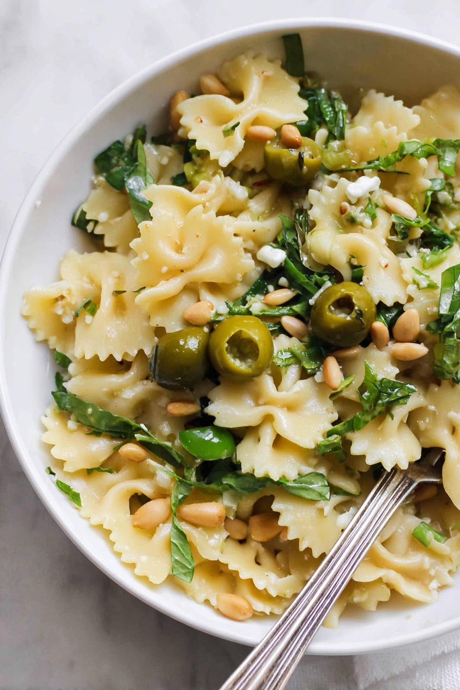 Dirty Martini Pasta Salad, easy pasta salad recipes, flavorful pasta salad ideas, briny pasta salad, party pasta salads - A close-up view of a white bowl filled with one layer of wavy-edged pasta pieces that are light yellow in color. Scattered on top are halved green olives, fresh green leafy herbs, and a few light brown pine nuts. There are also small pieces of a creamy white ingredient and some garlic cloves. A silver fork rests inside the bowl at the bottom right corner. The bowl is placed on a white marbled surface. Photo taken with an iphone --ar 2:3 --v 7