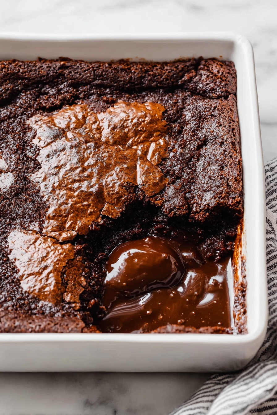 Decadent Hot Fudge Cake, hot fudge cake recipe, gooey chocolate cake, easy chocolate dessert, indulgent chocolate cake - A square white ceramic dish holds a rich chocolate dessert with a cracked, shiny top layer that is dark brown and slightly rough in texture. One section near the front corner is scooped out, revealing a thick, glossy, melted chocolate layer beneath the top crust. The melted chocolate is smooth and dark, contrasting with the more solid and matte top layer. The dish sits on a white marbled surface, and a striped cloth partially visible under one edge. photo taken with an iphone --ar 2:3 --v 7