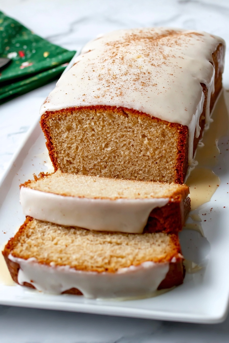 Eggnog Bread with Icing, holiday eggnog bread, festive bread recipes, Christmas flavored bread, easy eggnog dessert bread - A loaf cake with a light brown crust sits on a white rectangular plate against a white marbled surface. The cake is sliced twice, with two visible slices lying flat next to the main loaf. A thick white glaze covers the top of the cake, dripping over the sides and partly onto the plate, with a light dusting of cinnamon on top. The inside of the cake is a soft beige color with a slightly crumbly texture. Photo taken with an iphone --ar 2:3 --v 7