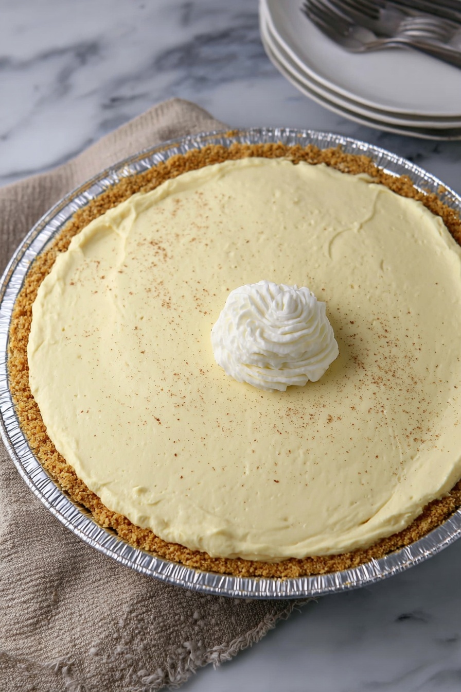 No Bake Eggnog Pie, Eggnog Pie, Holiday No Bake Desserts, Festive Eggnog Dessert, Easy Christmas Pie - A creamy light yellow pie with one slice missing is in a shiny round aluminum pan. It has two layers: a thick light brown crumb crust at the bottom and a thick smooth creamy yellow filling on top. The pie is topped with a small swirl of white whipped cream in the center and sprinkled with fine brown powder all over. The pie sits on a light brown woven cloth with a white marbled surface showing at the edge. On the right side, a partial view of stacked white plates with a fork is visible. photo taken with an iphone --ar 2:3 --v 7