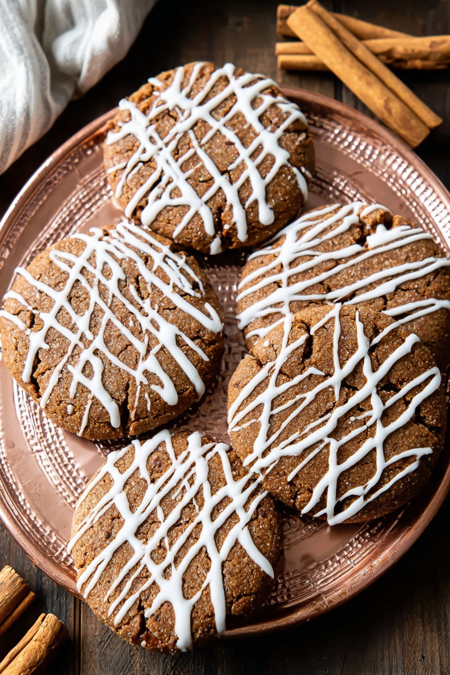 Soft Gingerbread Cookies, holiday gingerbread cookies, chewy gingerbread cookies, spiced gingerbread cookies, easy gingerbread cookies - A white plate holds eight round, light-brown cookies with a slightly cracked texture, each drizzled with thin white icing in a random, striped pattern across the tops. The cookies are evenly spaced on the plate, showing a soft and slightly raised surface with darker spots that hint at spices or chocolate bits. The plate sits on a white marbled surface, with a cinnamon stick and a pink cloth nearby, adding warm tones to the scene. Photo taken with an iphone --ar 2:3 --v 7