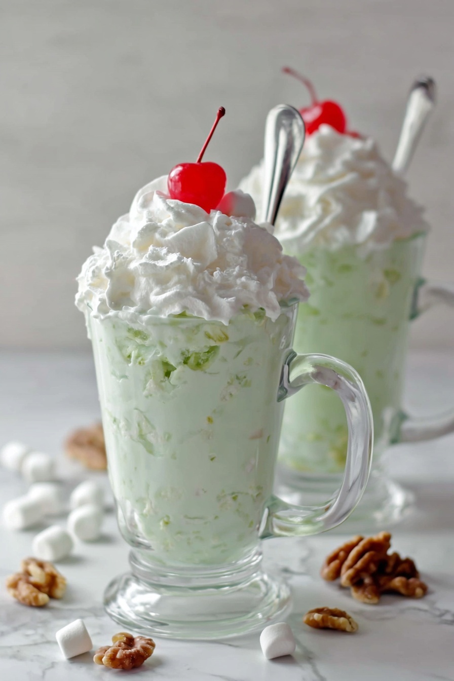 Easy Watergate Salad with Pineapple and Pecans, Watergate Salad recipe, quick fruity dessert, no-cook pineapple salad, nostalgic party side dish - Two clear glass mugs sit on a white marbled surface, each filled with a layered dessert. The bottom layer is a thick, creamy pale green mixture with visible small chunks, topped generously with a smooth white whipped cream layer. On the very top of each mound of whipped cream is a small, bright red cherry. Each mug has a long silver spoon inserted, leaning outwards. Around the mugs on the surface are scattered mini white marshmallows and small pieces of walnut. The photo taken with an iphone --ar 2:3 --v 7