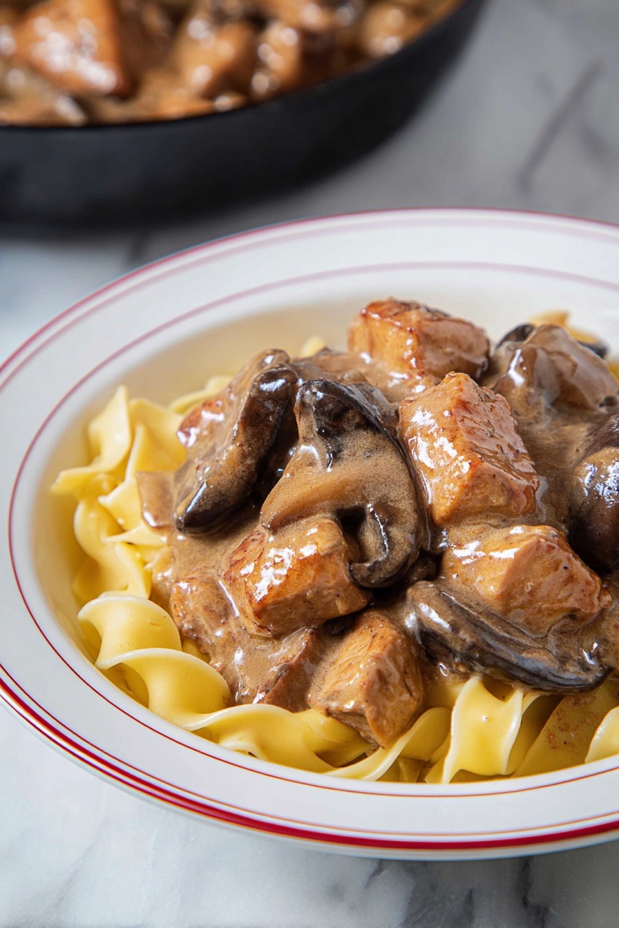 Easy Chicken Stroganoff, Chicken Stroganoff recipe, creamy chicken dinner, quick weeknight chicken, comforting chicken dish - The dish shows a layer of light yellow egg noodles at the bottom, with soft wide ribbons that curl slightly. On top, there is a thick layer of brown creamy sauce that coats chunks of cooked meat and slices of mushrooms. The meat pieces are uneven in size and have seared brown edges. The mushrooms are dark brown and smooth. Everything is served in a white bowl with a thin red ring around the edge. The bowl sits on a white marbled surface. In the background, there is a hint of a black skillet with more meat and sauce inside. Photo taken with an iphone --ar 2:3 --v 7