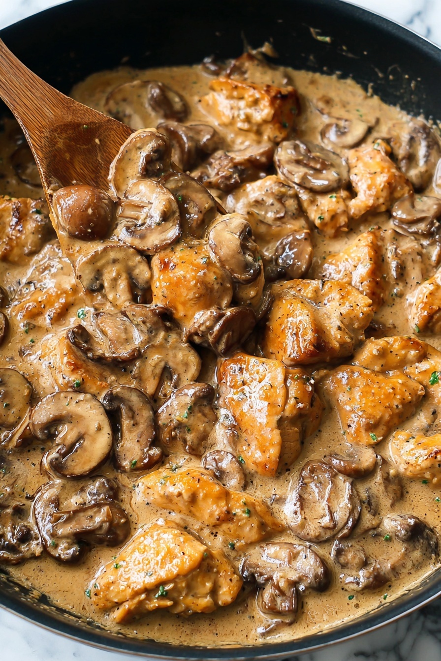 Easy Chicken Stroganoff, Chicken Stroganoff recipe, creamy chicken dinner, quick weeknight chicken, comforting chicken dish - The image shows a close-up of a pan filled with a creamy mushroom and chicken dish. The dish has two main layers: chunks of golden-brown chicken pieces and sliced brown mushrooms, both coated in a thick, light brown cream sauce. The chicken pieces are irregular in shape with a slightly crispy texture, while the mushrooms are soft and mixed throughout. A wooden spoon is partially submerged in the center, lifting some chicken and mushrooms. The pan is black and the background is a white marbled texture. Photo taken with an iphone --ar 2:3 --v 7