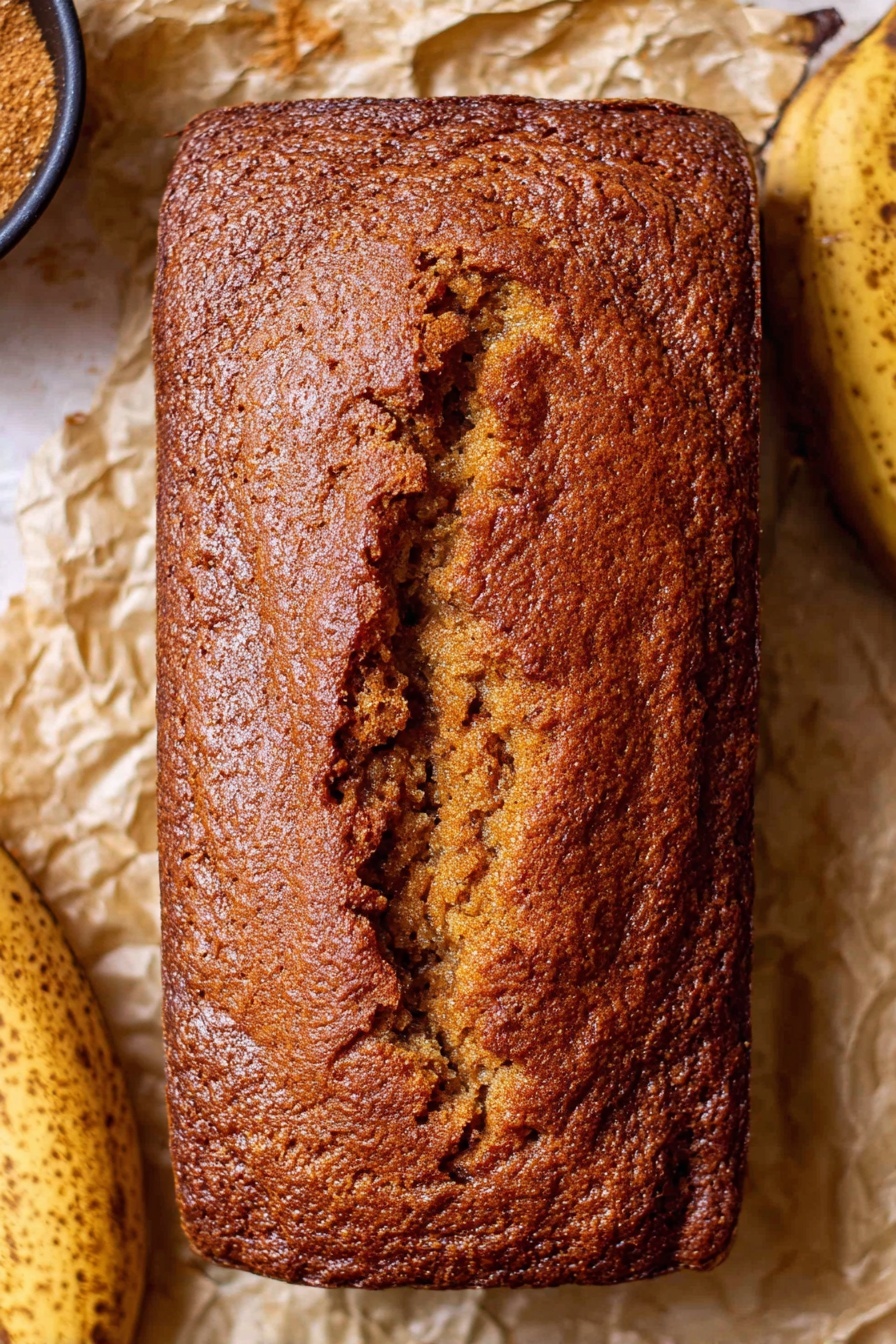 Moist Banana Bread, Banana Bread Recipe, Easy Banana Bread, Best Banana Bread, Homemade Banana Bread - A single loaf of brown banana bread with a rough and cracked crust texture, sitting directly on crumpled parchment paper. The bread is rectangular with rounded corners and a deep crack running lengthwise down the middle. To the sides of the bread are parts of ripe bananas with dark spots, and part of a small black bowl with brown sugar is slightly visible on the left. The background is a white marbled texture. Photo taken with an iphone --ar 2:3 --v 7