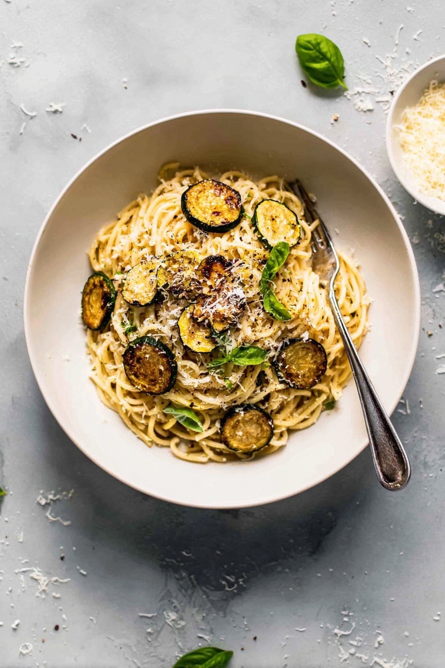 Creamy Zucchini Spaghetti, zucchini pasta with creamy sauce, summer zucchini recipes, easy zucchini pasta, healthy zucchini spaghetti - A white bowl filled with a mound of spaghetti pasta at the center, topped with several golden-brown, round slices of roasted zucchini scattered on and around the noodles. Small bright green basil leaves are placed over the pasta, adding a fresh touch. The spaghetti is sprinkled with finely grated white cheese, giving a light dusting over the top. A silver fork rests on the right side inside the bowl. The bowl is set on a white marbled surface with some scattered grated cheese and a few basil leaves nearby. Photo taken with an iphone --ar 2:3 --v 7