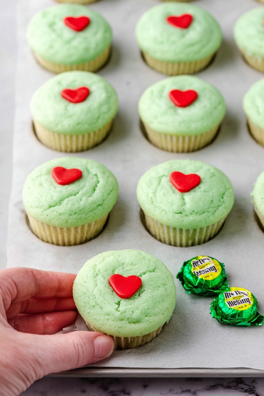 Green Hershey’s Kiss Cookie Cups, festive holiday cookie recipes, easy chocolate cookie bites, no-bake cookie cups, green candy cookie treats - A dark gray muffin tray holds 18 small round green cookies inside each cup, with a soft and slightly bumpy texture. A woman's hand with green nail polish is placing a small red heart-shaped decoration on one of the cookies in the center. Some cookies already have a red heart decoration pressed slightly into their surface. The tray rests on a white marbled surface. photo taken with an iphone --ar 2:3 --v 7