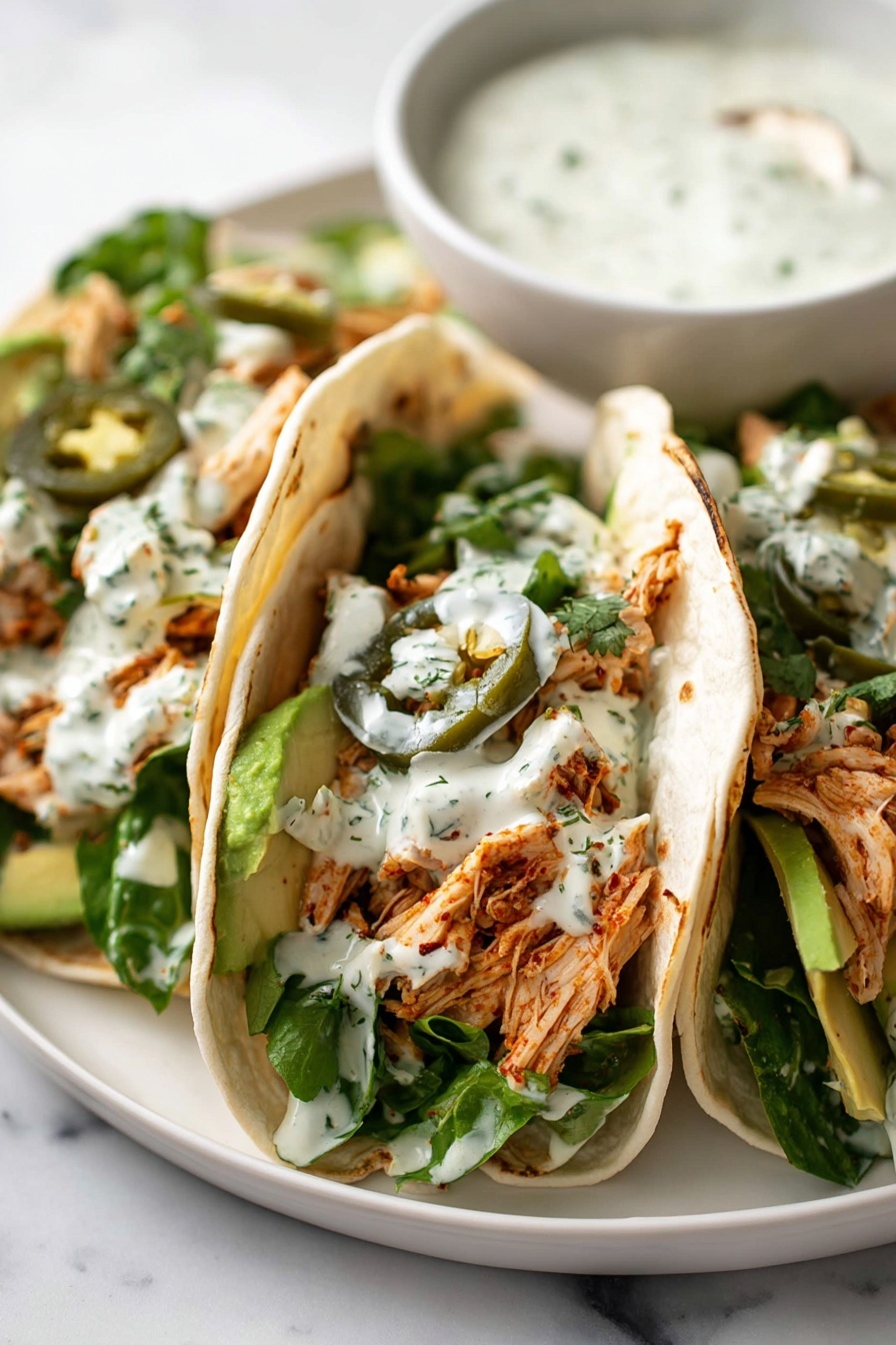 Shredded Chicken Tacos with Creamy Chimichurri, chicken taco ideas, easy chicken tacos, fresh chimichurri sauce, flavorful taco recipes - The image shows three soft white tortillas folded in half and filled with shredded seasoned chicken, green leafy lettuce, thin slices of avocado at the bottom, and sliced jalapeños scattered on top. There is a creamy white sauce with herbs drizzled over the filling. The tacos are placed closely together on a white plate with a white marbled surface underneath. In the background, there is a white bowl filled with the same creamy sauce. photo taken with an iphone --ar 2:3 --v 7