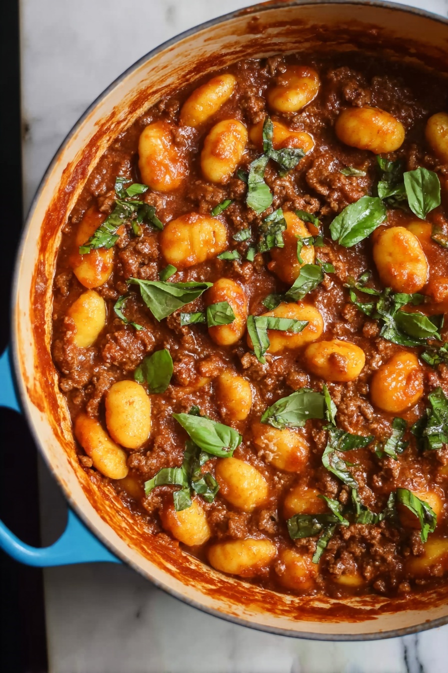 Gnocchi Bolognese, easy gnocchi bolognese recipe, hearty gnocchi dinner, homemade bolognese sauce, comforting pasta dishes - The image shows a blue pot filled with a rich brown meat sauce mixed with soft, yellow-orange gnocchi. The top layer is scattered with pieces of fresh green herbs, adding a bright contrast to the warm colors of the sauce and gnocchi. The pot's handle is visible on the right side, resting on a white marbled surface. The texture of the gnocchi looks smooth and tender, while the meat sauce appears thick and hearty, covering all pieces evenly. Photo taken with an iphone --ar 2:3 --v 7