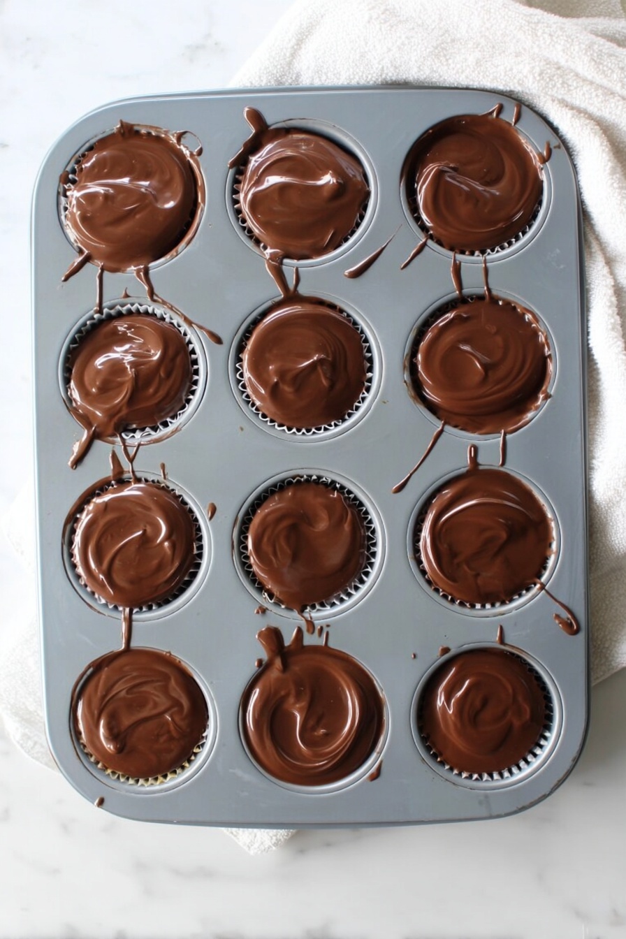 No-Bake Peanut Butter Oat Cups, healthy no-bake snack, quick no-bake dessert, easy peanut butter snack, wholesome oat cups - A gray muffin tray with twelve round cups, each filled with a smooth, glossy layer of melted chocolate that slightly spills over the edges. The chocolate has a shiny, swirled texture on top, and thin chocolate drips and streaks connect some of the cups and run down the tray’s sides. The tray rests on a white marbled surface with a soft white cloth partially visible at the top right corner. Photo taken with an iphone --ar 2:3 --v 7