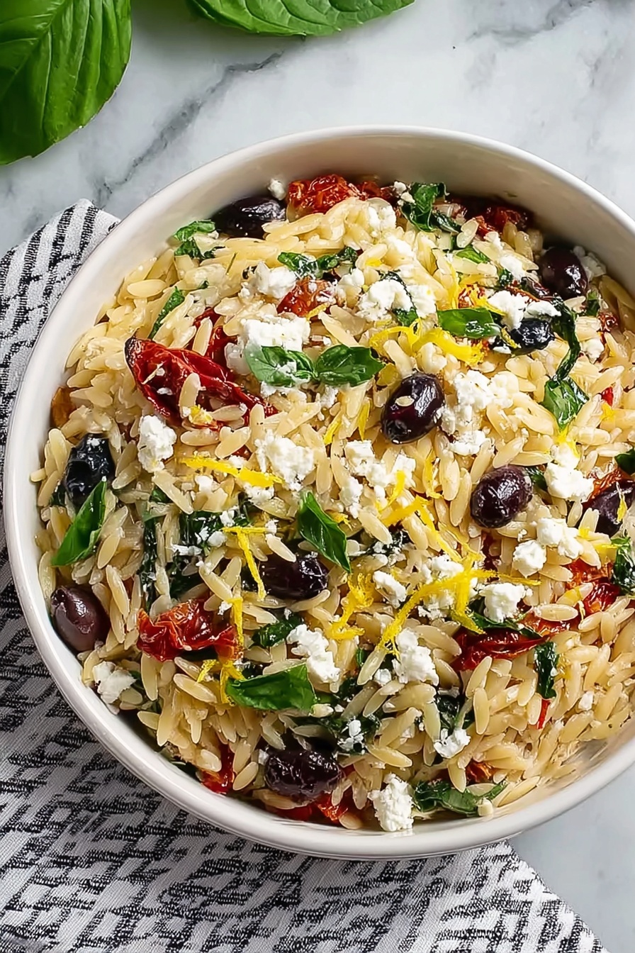 Orzo Pasta Salad with Feta and Sun-Dried Tomatoes, Mediterranean pasta salad, easy pasta salad recipes, feta pasta salad, sun-dried tomato pasta - A round white bowl filled with a layered dish placed on a striped cloth over a white marbled surface. The bottom layer consists of small, light golden orzo pasta. Mixed into the orzo are dark purple olives, green fresh basil leaves, and pieces of sun-dried red tomatoes. On top, crumbled white feta cheese is scattered evenly, along with small bright yellow lemon zest strips adding color contrast. The textures vary from soft pasta, crumbly cheese, to smooth olives and delicate leaves. Photo taken with an iphone --ar 2:3 --v 7