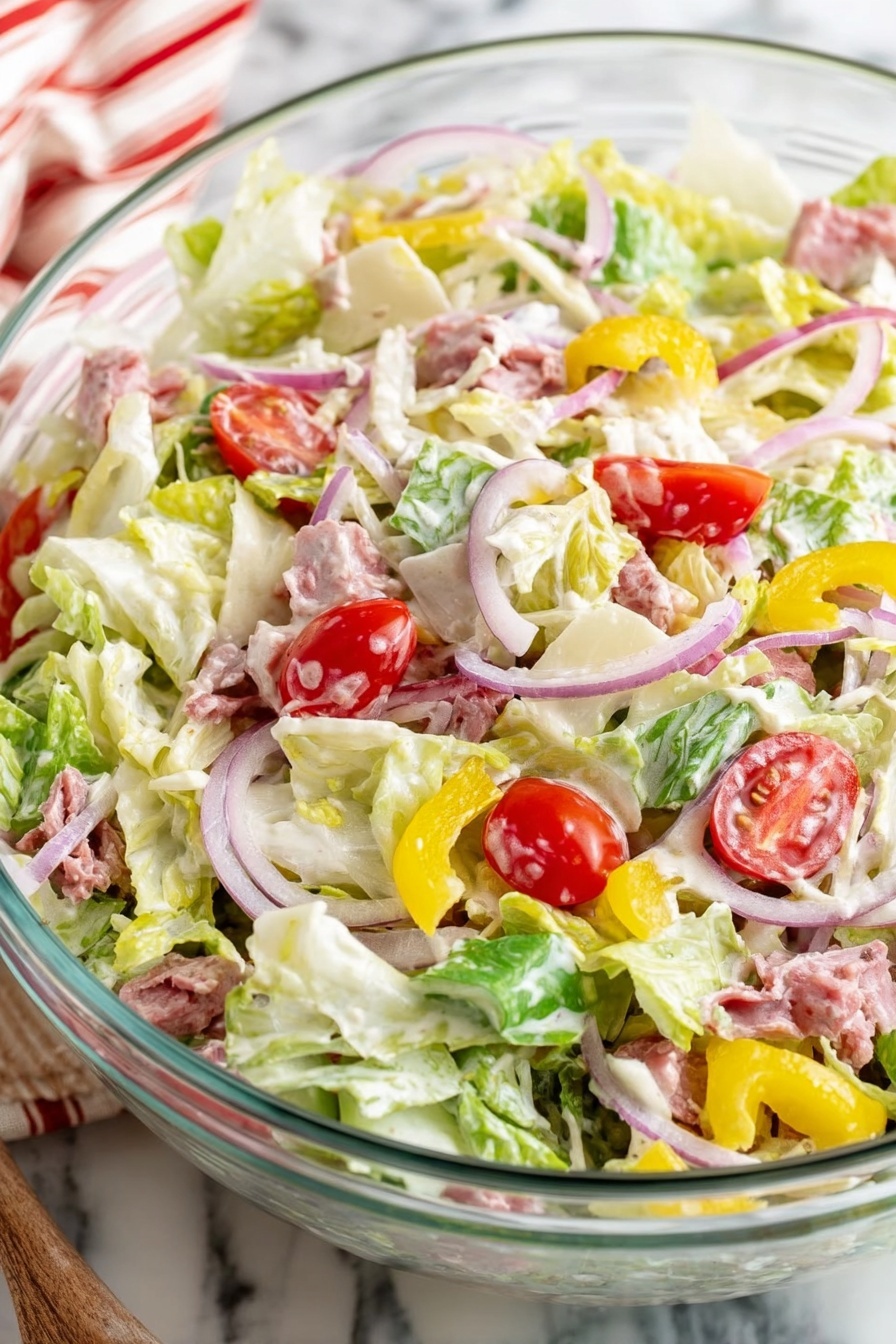 Grinder Salad with Parmesan and Salami, hearty Italian salad, quick flavorful salad, salami and Parmesan salad, easy salami salad recipe - A clear glass bowl filled with a fresh salad showing three main layers: the base layer is mostly light green and pale yellow chopped lettuce with some creamy dressing coating it; scattered on top are slices of red cherry tomatoes, thin rings of purple onion, yellow pepper pieces, and small chunks of pale cheese; mixed throughout are small pieces of meat that are pinkish in color. The bowl sits on a white marbled surface with part of a red and white striped cloth visible in the background photo taken with an iphone --ar 2:3 --v 7