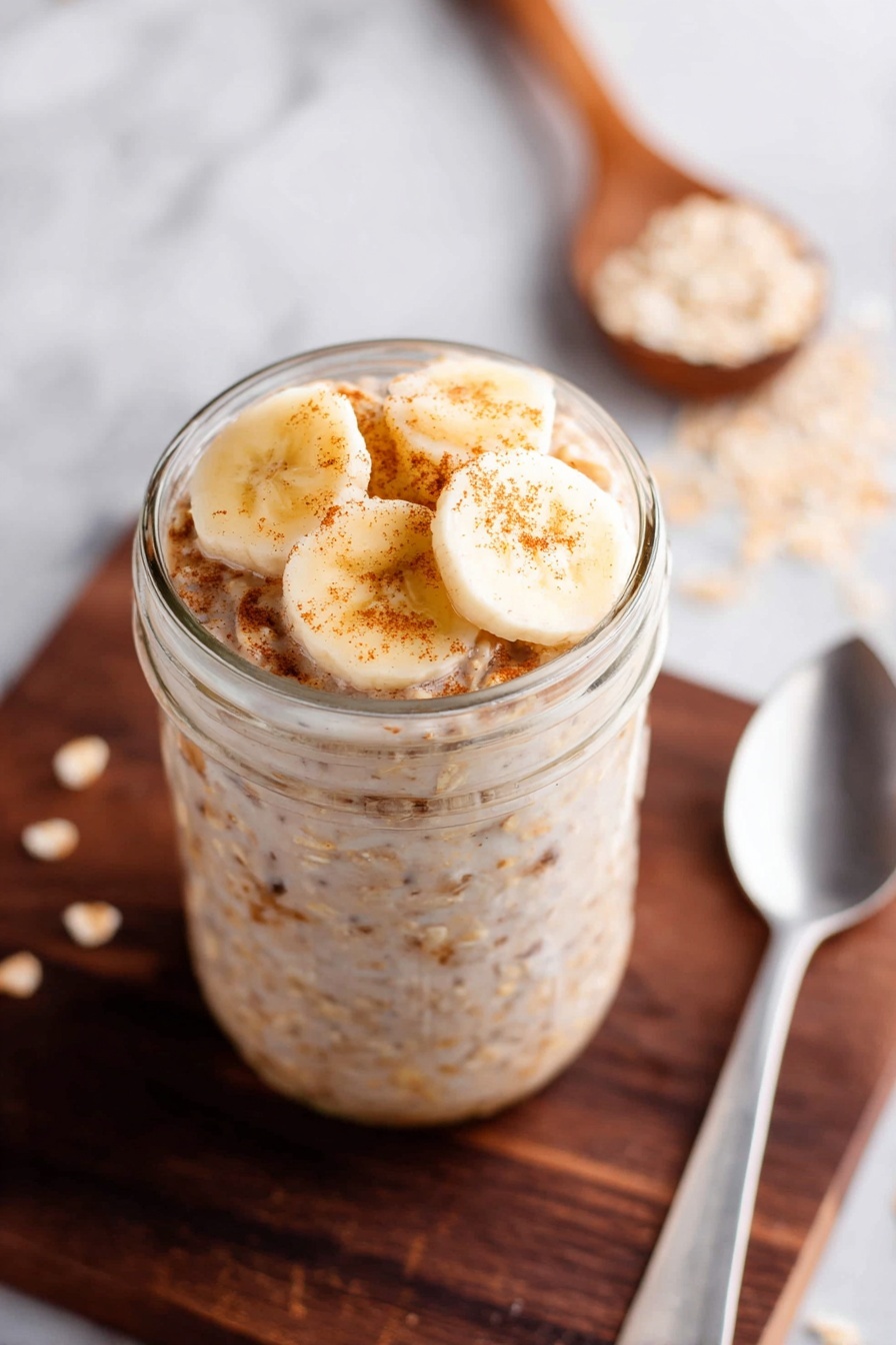 Peanut Butter Banana Overnight Oats, healthy breakfast ideas, easy overnight oats, nutritious morning recipes, quick make-ahead breakfast - A clear glass jar with a silver metal lid filled with three layers of overnight oats. The bottom layer is creamy white with tiny pieces of oats and seeds showing through. The middle layer has a light brown chunk inside the creamy oats mixture creating a rough texture. The top layer blends smoothly into the silver lid. The jar sits on a white marbled surface with scattered oats around, a white cloth on the left, and a yellow banana on the right photo taken with an iphone --ar 2:3 --v 7