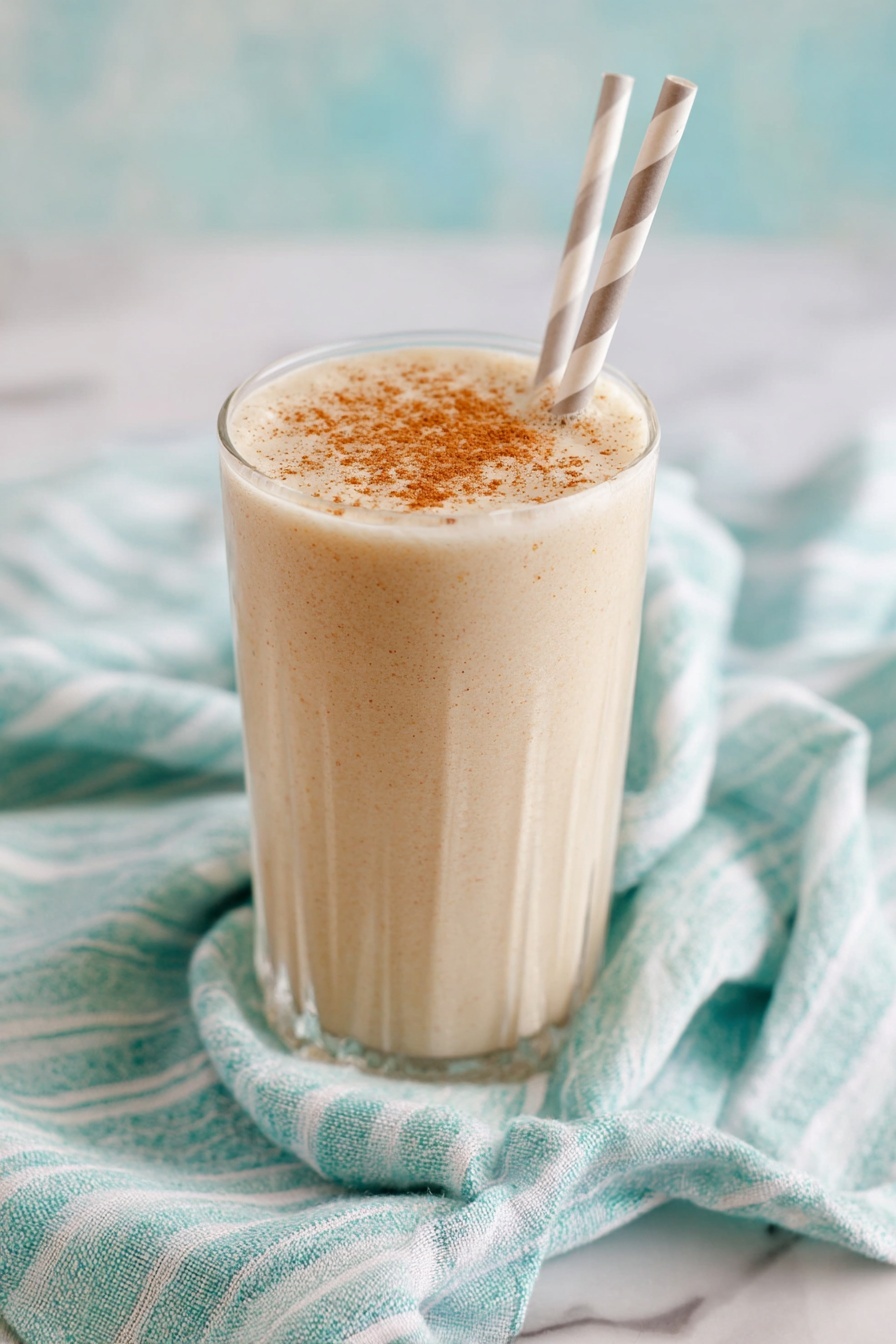 Vanilla Chai Protein Shake, Vanilla Chai Protein Smoothie, Healthy Chai Protein Drink, Protein Shake with Spices, Cozy Morning Protein Shake - A tall clear glass filled with a creamy, light beige drink that has small specks mixed throughout, showing a smooth and thick texture. The top of the drink is sprinkled evenly with a light brown powder. The glass sits on a green and white striped cloth on a white marbled surface. The background is soft and out of focus with light creamy tones. photo taken with an iphone --ar 2:3 --v 7