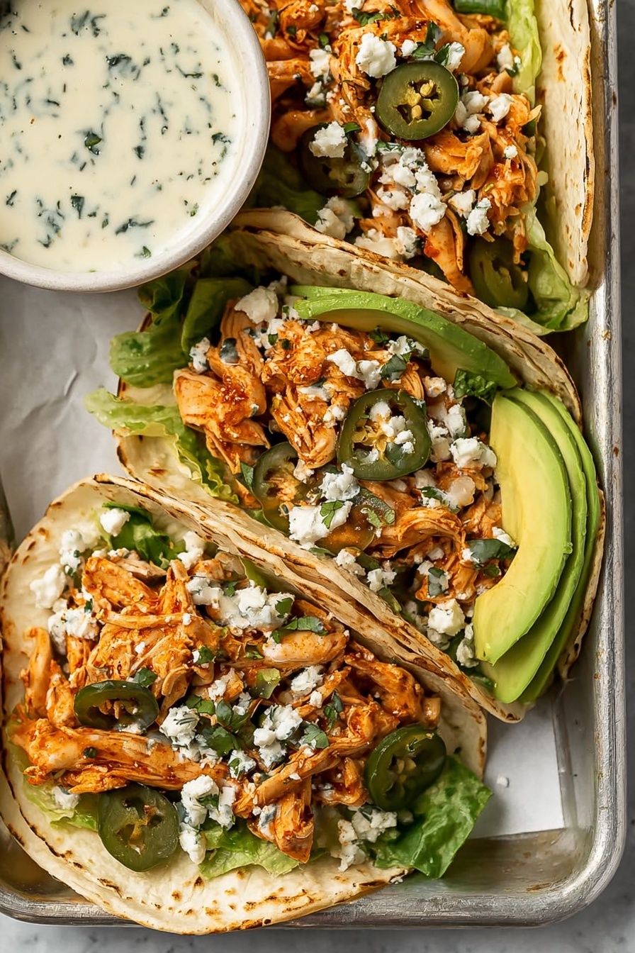 Shredded Chicken Tacos with Creamy Chimichurri, chicken taco ideas, easy chicken tacos, fresh chimichurri sauce, flavorful taco recipes - Three white soft tacos are placed closely on a metal tray with a white marbled surface underneath. Each taco has a layer of light green lettuce at the bottom, followed by orange shredded chicken mixed with bits of red onion and dark green sliced jalapeños. On top of the chicken is a sprinkle of white crumbly cheese. Thin slices of bright green avocado are tucked on the side of the tacos. In the top left corner of the tray, a white bowl holds a creamy sauce with small green herb pieces visible. The overall look is fresh with soft, layered textures. photo taken with an iphone --ar 2:3 --v 7